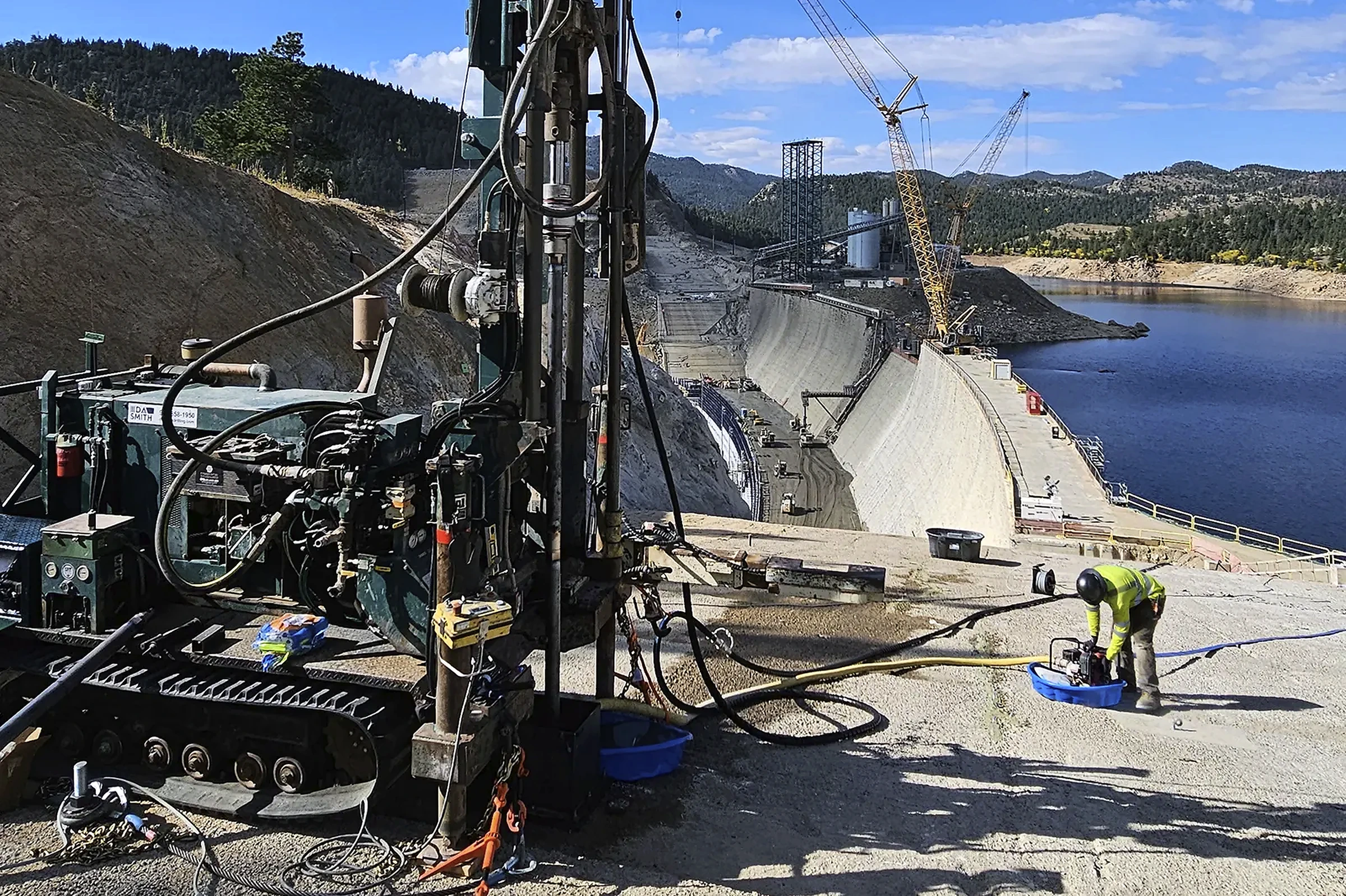 Drilling equipment and crew beside a dam and reservoir